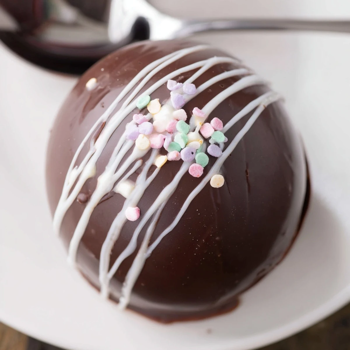 Vivid red-sprinkled Hot Chocolate Bombs sit on a white plate next to a steaming mug.
