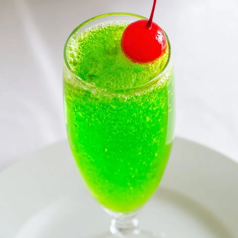 Bright green Quick Grinch Mimosa Mocktail, served in a champagne flute, garnished with a cherry.