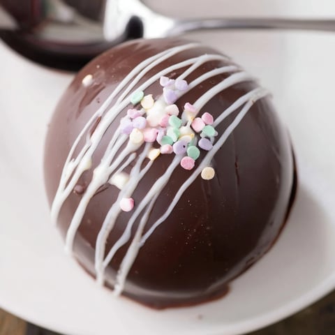 Vivid red-sprinkled Hot Chocolate Bombs sit on a white plate next to a steaming mug.