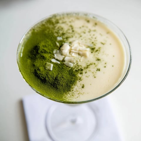A vibrant Matcha Martini cocktail in a chilled glass, garnished with matcha powder dusting beside small white chocolate shavings.  