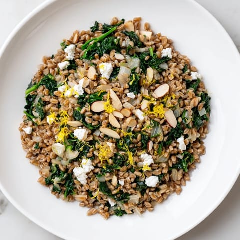 Close-up of a steaming bowl of Warm Farro & Greens Pilaf sprinkled with toasted almonds and feta.