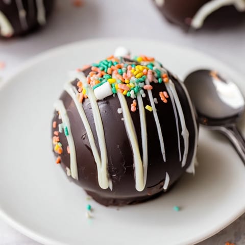 A hand holding a spoon cracks open a glossy Hot Chocolate Bomb as marshmallows spill out.
