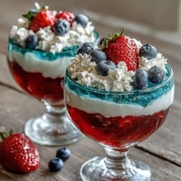 Red, White, and Blue Layered Jello Cups with vibrant stripes, topped with whipped cream and fresh berries for a patriotic treat.